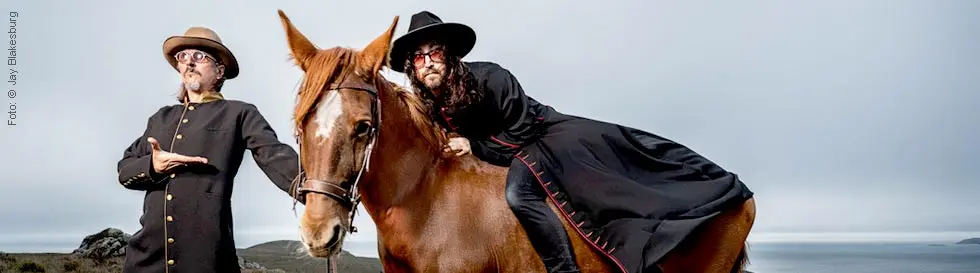 Zwei Personen in schwarzen Gewändern posieren neben und auf einem braunen Pferd; sie tragen Hüte und Sonnenbrillen.