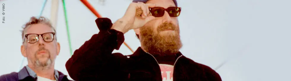 Links ein Mann mit Brille, rechts ein Mann mit Bart, Sonnenbrille und Mütze. Hintergrund zeigt ein farbiges Karussell.