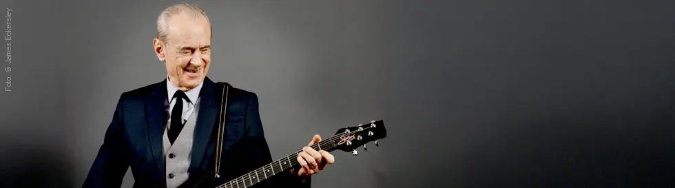 Ein älterer Mann im Anzug spielt lächelnd eine E-Gitarre vor einem grauen Hintergrund.