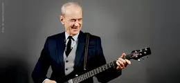 Ein älterer Mann im Anzug spielt lächelnd eine E-Gitarre vor einem grauen Hintergrund.