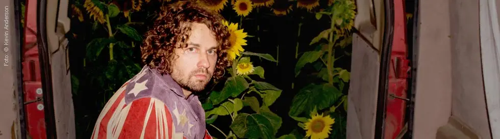 Mann mit lockigen Haaren, in bunter Fransenjacke, sitzt in geöffnetem Van. Im Hintergrund leuchtende Sonnenblumen.