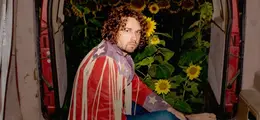 Mann mit lockigen Haaren, in bunter Fransenjacke, sitzt in geöffnetem Van. Im Hintergrund leuchtende Sonnenblumen.