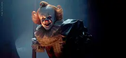 Clown mit weiß geschminktem Gesicht, roter Nase und orangefarbenem Haar, lächelt im Dunkeln bedrohlich.