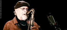 Ein Mann mit Brille und Mütze spielt Saxofon vor einem Mikrofon. Dunkler Hintergrund.