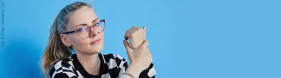 Eine Person mit Brille, gestreiftem Pulli sitzt vor hellblauer Wand, hält eine Hand in der anderen.