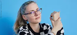Eine Person mit Brille, gestreiftem Pulli sitzt vor hellblauer Wand, hält eine Hand in der anderen.