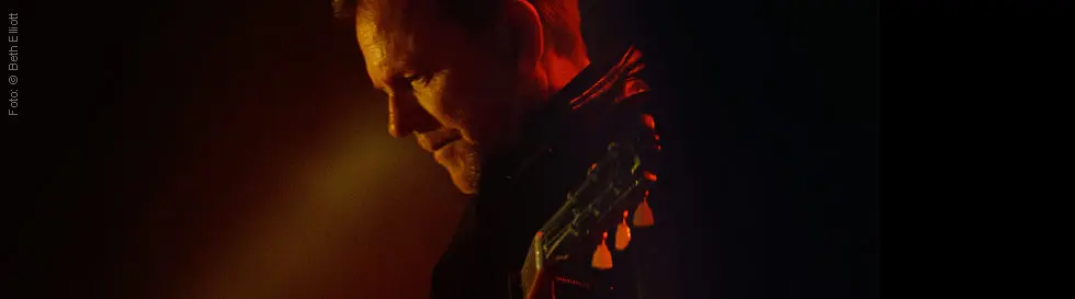 Ein Mann mit einer Gitarre, in rotes Licht getaucht, schaut nach unten.