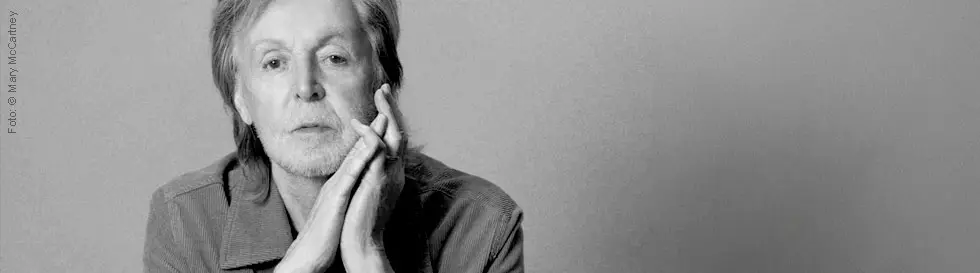 Paul McCartney betrachtet dich nachdenklich, beide Hände am Kinn, grauhaarig.