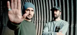 Zwei Männer in einem Tunnel, einer hebt die Hand zur Kamera, der andere steht mit verschränkten Armen.