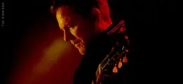 Ein Mann mit einer Gitarre, in rotes Licht getaucht, schaut nach unten.