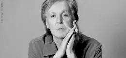 Paul McCartney betrachtet dich nachdenklich, beide Hände am Kinn, grauhaarig.