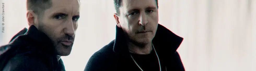Zwei Männer in dunkler Kleidung vor einem hellen Hintergrund, ernst blickend. Einer trägt eine Kette, beide haben kurzes Haar.