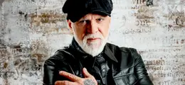 Eine Person mit weißem Bart und schwarzer Mütze, trägt eine schwarze Lederjacke, Hintergrund: Verblasste Buchstaben auf einer hellen Wand.