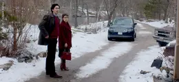 Zwei Personen stehen auf einem schneebedeckten Weg, links ein Mann, rechts eine Frau mit rotem Mantel, im Hintergrund zwei Autos.