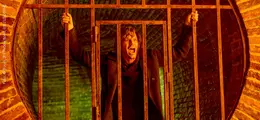 Eine Person schreit hinter einem Metallgitter in einem Tunnel mit gemauerten, orange-beleuchteten Wänden.