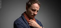 Eine nachdenkliche Person mit braunem Schal und blauem Mantel schaut nach unten, vor einem neutralen Hintergrund.