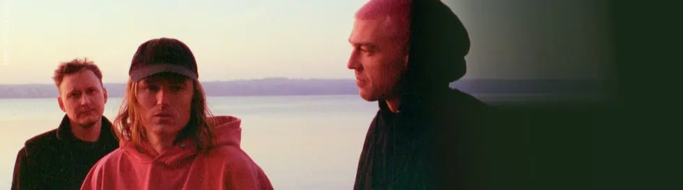Drei Männer stehen am Wasser. Einer trägt eine rosa Kapuzenjacke und eine Mütze, die anderen dunkel gekleidet in Schwarz.