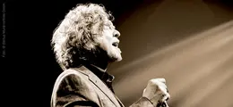 Ein Mann mit lockigem Haar singt leidenschaftlich in ein Mikrofon, dramatisch beleuchtet mit einem Spotlicht von der Seite.