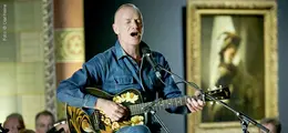 Ein Mann spielt Gitarre und singt. Im Hintergrund hängt ein Gemälde in einem goldenen Rahmen.