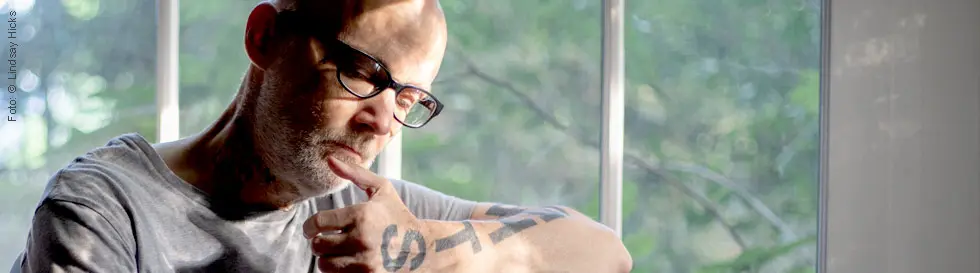 Ein Mann mit Brille und Tattoos sitzt nachdenklich am Klavier. Hinter ihm große Fenster mit Blick ins Grüne.