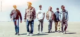 Sechs Männer gehen lässig über einen Strand, in casual Kleidung und Sonnenbrillen, bei sonnigem Wetter.