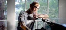 Ein Mann mit Brille und Tattoos sitzt nachdenklich am Klavier. Hinter ihm große Fenster mit Blick ins Grüne.