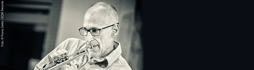 Eine schwarz-weiß Fotografie zeigt einen älteren Mann mit Brille, der konzentriert Trompete spielt.