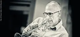 Eine schwarz-weiß Fotografie zeigt einen älteren Mann mit Brille, der konzentriert Trompete spielt.