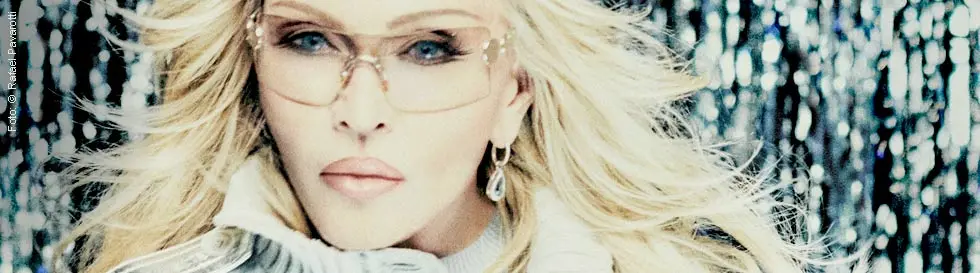 Eine Person mit langen, blonden Haaren trägt eine Brille und silberne Kleidung.