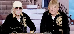 Zwei Männer in schwarzen Hemden mit goldenen Mustern. Einer trägt Sonnenbrille und Mütze, beide haben eine Gitarre.