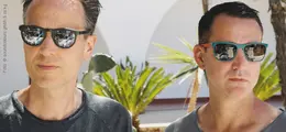 Zwei Männer mit Sonnenbrillen stehen draußen, umgeben von Pflanzen und Licht. Sie schauen ernst in die Ferne.