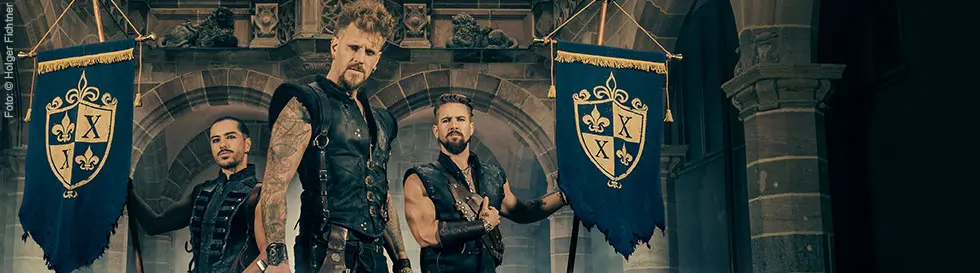 Zwei blaue Banner mit goldenen Wappen, jeweils mit einem "X" und einer Lilie. Drei Männer in Lederkleidung posieren.