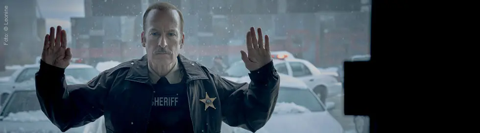 Ein Mann im Sheriff-Outfit hebt die Hände, steht vor schneebedeckten Autos, deren Lichter leuchten, mit ernster Miene.