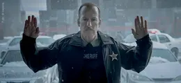 Ein Mann im Sheriff-Outfit hebt die Hände, steht vor schneebedeckten Autos, deren Lichter leuchten, mit ernster Miene.