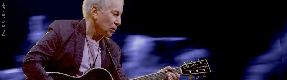 Ein älterer Mann spielt Gitarre vor einem unscharfen, dunklen Hintergrund.