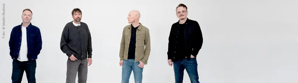 Vier Männer in lässiger Kleidung stehen in einer Reihe vor einem hellen Hintergrund.