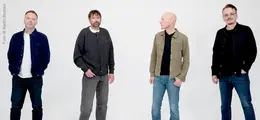 Vier Männer in lässiger Kleidung stehen in einer Reihe vor einem hellen Hintergrund.
