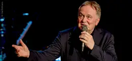 Ein Mann mit Mikrofon singt gestikulierend auf einer Bühne in dunkler Kleidung.