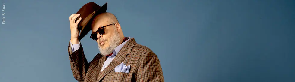 Ein Mann mit Sonnenbrille und Hut, gekleidet im karierten Anzug, hebt den Hut leicht an, vor einem blauen Hintergrund.