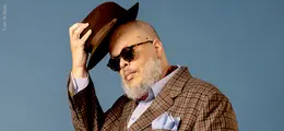 Ein Mann mit Sonnenbrille und Hut, gekleidet im karierten Anzug, hebt den Hut leicht an, vor einem blauen Hintergrund.