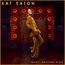 "KAT EATON; WHAT HAPPENS NOW." Eine Frau in einem goldfarbenen Anzug steht vor einer Wand mit orangefarbenen Lautsprechern.