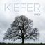 "Kiefer Sutherland, GREY" in großen Buchstaben. Ein kahler Baum unter grauem Himmel, ruhig und imposant.