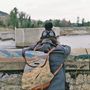 Text auf dem Rucksack: Ein Pfad durch Landschaft. Person mit Wollmütze blickt auf einen Fluss hinter einer Steinmauer., LP