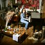 Eine Frau in Jeans spielt Klavier in einem vollen Raum, mit Musikinstrumenten, Dekorationen und einem Hund auf Holzboden., LP