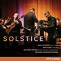 "Solstice" in großen Buchstaben. Musiker mit Gitarren und Bandoneon, Orchester im Hintergrund. Warmes Licht, Konzertatmosphäre., CD