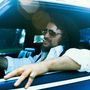 Ein Mann mit Hut und Sonnenbrille lehnt lässig im Auto, eine Zigarette im Mund.