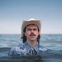 Mann mit Cowboyhut und Schnurrbart steht im Wasser, trägt ein blaues Hemd. Im Hintergrund ist der Horizont sichtbar., LP