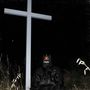 Jemand in schwarzer Kleidung vor einem hohen Kreuz. Sie tragen eine Maske mit weißen Schriftzeichen und stehen im Gras., LP
