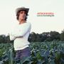 „ARTHUR RUSSELL Love Is Overtaking Me“. Eine Person mit Hut steht in einem Feld, umgeben von Pflanzen.