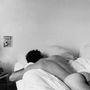 Ein schwarz-weißes Foto zeigt eine Person, nackt auf dem Bauch in einem Bett liegend, in einem spartanischen Raum mit einem Poster an der Wand., LP